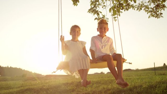SLOW MOTION CLOSE UP: Young brother and sister swinging on swing under a tree on warm summer evening. Little boy and girl enjoying golden sunset on a swing. Happy kids swinging outdoors in summer.