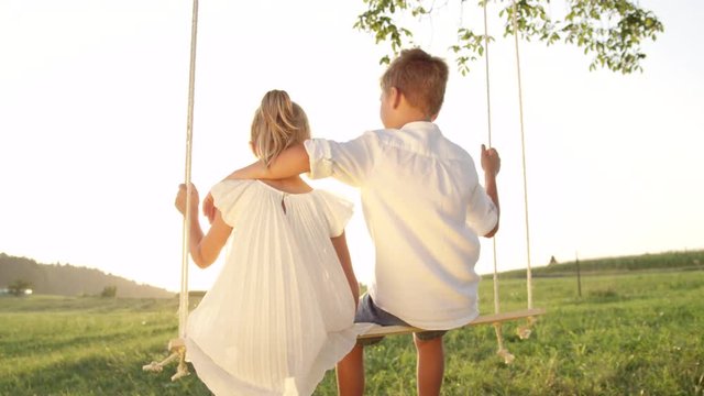 SLOW MOTION CLOSE UP: Couple of happy kids hugging and swinging on wooden swing at golden sunset. Brother embracing little sister on a swing. Young love between boy and girl on romantic summer evening
