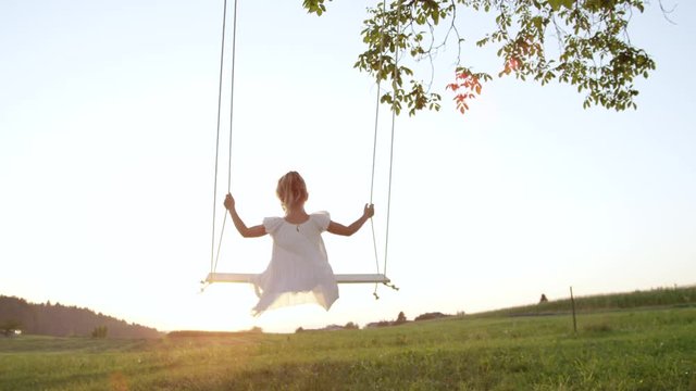 SLOW MOTION CLOSE UP Unrecognizable little girl swaying on a wooden swing on a warm summer evening. Girl in a flowing white dress enjoying sunset. Barefoot child on a swing looking at the setting sun.