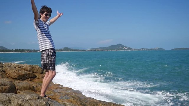 Successful happy businessman raising hands to the sky by the sea with splashing waves on the rock in slow motion. 1920x1080