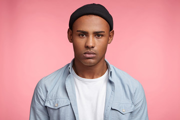 Obraz premium Serious fashionable male with dark skin, wears black hat and casual shirt, looks directly into camera, has serious expression, isolated over pink background. People, youth, style, facial expressions