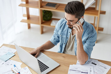 Bearded skilled young talented copywriter works on new article, keyboards at laptop, has phone...