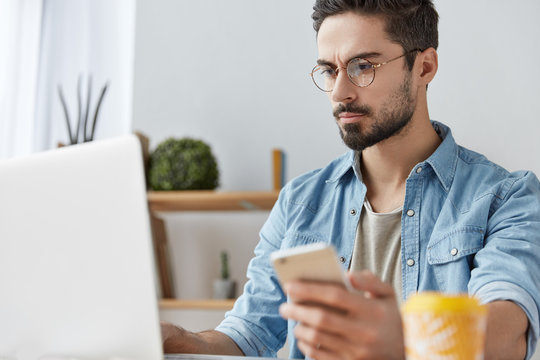 Portrait of handsome bearded male freelancer installs new application on smart phone, downloads program at laptop computer, uses wireless internet connection, recieves email or message from partner