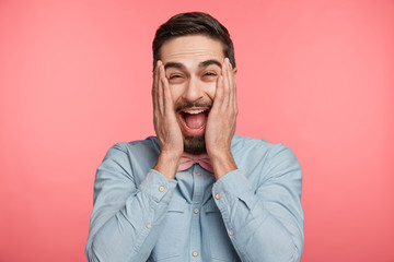 Funny overjoyed glad cheerful bearded male dressed formally, laughs at good joke, can`t stop giggling, keeps hands on cheeks, keeps mouth widely opened, expresses positiveness and happiness.