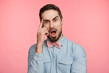 People, facial expressions and emotions concept. Shocked amazed young male with trendy hairdo, wears formal clothes, keeps index finger on temple, keeps mouth widely opened, being emotional.