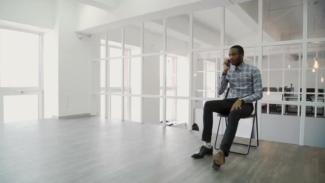 Young businessman talking on phone while sitting in modern office. African man makes important call with partner, using a smartphone. Successful rustler dressed in stylish clothes with watch on wrist