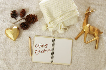 Notebook with text: MERRY CHRISTMAS and cup of cappuccino over cozy and warm fur carpet. Top view.