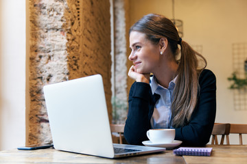 young attractive woman posing for a portrait sitting at a table in a cafe with a satisfied expression. Coffee mug, laptop help it to achieve success in business