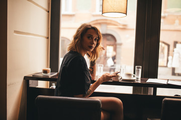 young adolescent woman in her late twenties, sitting in a bar at the window, drinking coffee and talking to a friend on a phone 