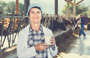 Female farmer is standing with glass of cow milk at the farm.