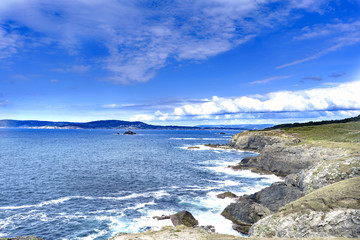 Rocky cliffs on the Atlantic coast of Galicia in a large bay and with a small island in the center. Calm sea and sky with some clouds