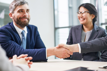 Portrait shot of confident entrepreneur sitting at table of modern boardroom and shaking hand of unrecognizable business partner as sign of successful completion of negotiations