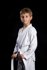 Smiling karate kid in brand new kimono, posing on the black background. Karate fighter holding his belt