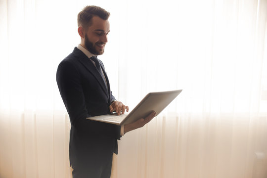 Side view of successful bearded businessman using laptop and smiling standing against background of window curtains in hotel room, copy space