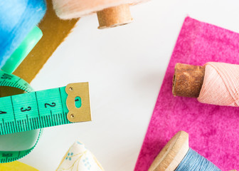 patchwork, sewing and tailoring concept - close-up on tools, measuring meter, blue and pink threads in bobbins and purple and mustard color patches on white background, top view, flat lay