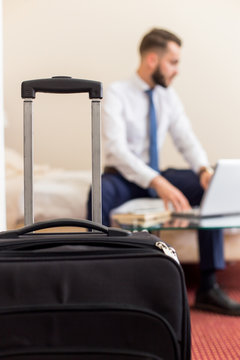 Blurred portrait of handsome bearded businessman using laptop working in hotel room, focus on suitcase in foreground