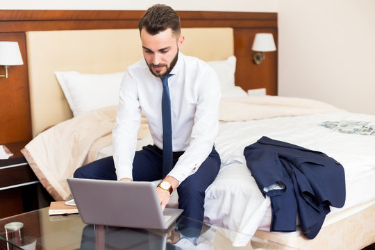 Portrait of handsome bearded businessman using laptop working in hotel room sitting on bed and enjoying business travel, copy space