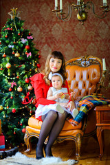 happy mother sit on a chair near the festive tree and hugs her little daughter with a small gift in her hands.