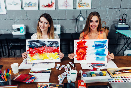Two smiling female illustrators working in their workshop holding paper sheets with flags of France and Germany drawn with watercolor technique