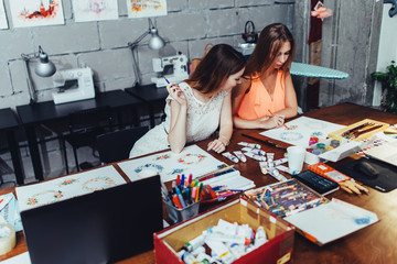 Two female adult students drawing pictures with crayons sitting at desk covered with painting...