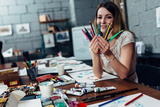 Smiling young woman holding a bunch of crayons sitting at desk surrounded by art supplies