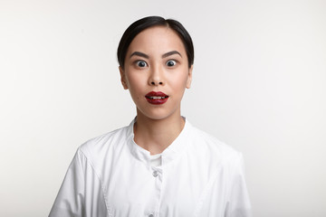 Picture of astonished bug eyed young Chinese woman cosmetologist in uniform expressing shock and surprise, raising eyebrows and opening mouth. Human facial expressions, emotions and feelings