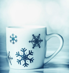 White mug with blue snowflakes on blue table and bokeh background, front view. Happy winter holiday card layout