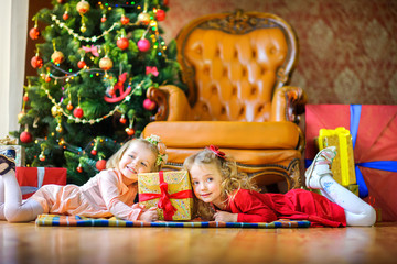 two girls are lying on the floor near gifts and smiling, in the background a festive Christmas tree.