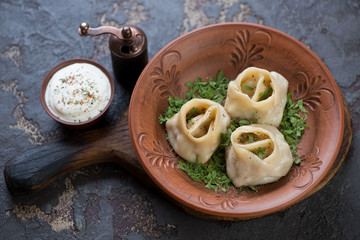 Clay plate with steamed meat dumplings or manti served with fresh greens and sour cream, brown stone background, horizontal shot