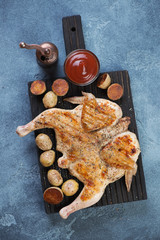 Top view of roasted chicken tabaka with cherry potato on a black wooden serving board, blue stone background, vertical shot