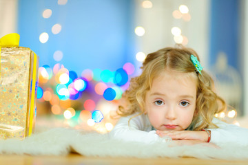 beautiful little girl lies on the floor on a white blanket, next is a gift, in the background festive lights.