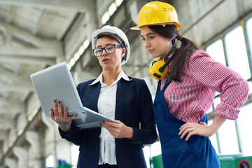 Pensive female building contractor demonstrating online sketch on portable computer to colleague in workwear in factory workshop