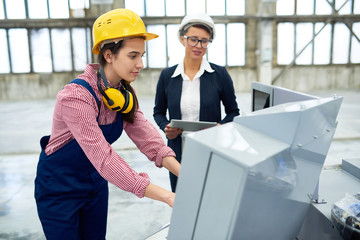 Confident female factory worker in hardhat wearing protective ear muffs on neck entering data in CNC machine while lady boss overseeing her work