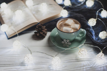 Cozy winter home. Cup of cocoa with marshmallows, warm knitted sweater, open book, Christmas garland, lump on a white wooden table. Atmosphere of a pleasant evening for reading.