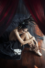 Asian ballerina woman nude sits on the floor and looking to white skeleton paper marche mask in the dark bed room natural window light.