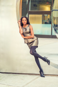 African American Business Woman Fashion in New York. Lady wearing metal crystal sleeveless mini dress top, skit suit, black leggings, high heels, leans against silver metal wall, relaxing, thinking..