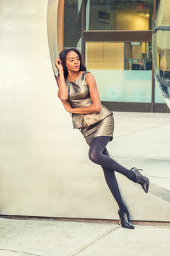 African American Business Woman Fashion in New York. Lady wearing metal crystal sleeveless mini dress top, skit suit, black leggings, high heels, leans against silver metal wall, relaxing, thinking..