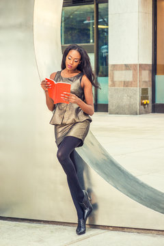 I love reading book. African American woman wearing metal crystal sleeveless mini dress top, skit suit, black leggings, high heels, holding red book, leans against silver metal wall, reads..