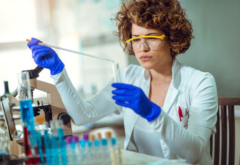Young female scientist in protective glasses and gloves preparing a liquid substance for test tube with a long glass pipette in the scientific chemical laboratory
