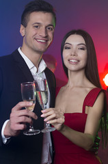 A group of young cheerful and happy friends are dancing at a party. A man and a woman are dancing in the foreground and clinking glasses with champagne