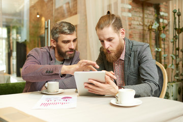 Team of hard-working managers using digital tablet while having project discussion at cozy coffeehouse with stylish interior