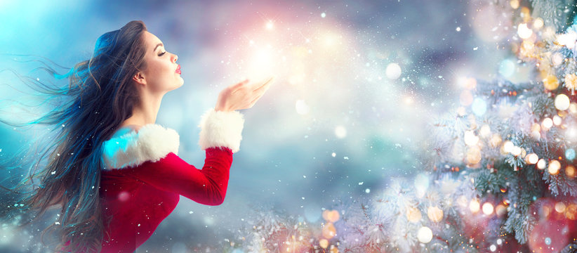 Christmas scene. Sexy Santa. Brunette young woman in party costume blowing snow over holiday blurred background