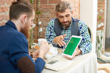Bearded middle-aged man wearing checked shirt and waistcoat showing statistic data to his male colleague with help of digital tablet while having informal working meeting at cafe.
