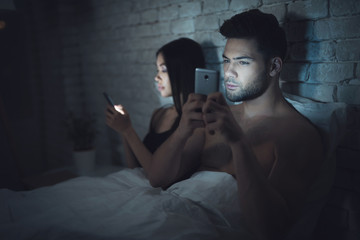 Girl with guy lies in a dark room in bed. They are looking at the smartphone. Influence of social networks.