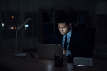 A man is sitting at a laptop in a dark office. The man in the suit works.