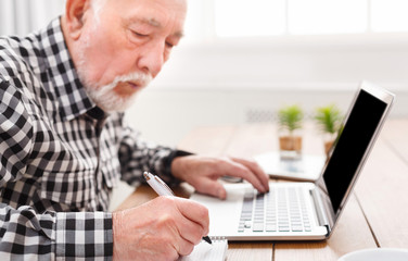 Mature man using laptop and writing in notepad