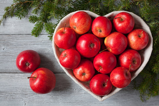 Red apples, basket in the shape of heart on a wooden background and spruce branches.