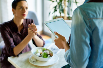 Close up of young cafe waitress holding digital tablet taking order from client sitting at table