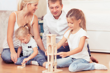 Cheerful family spending time together at home,. Daddy, mommy and little daughters lying on the wooden floor and playing with a wooden tower game.