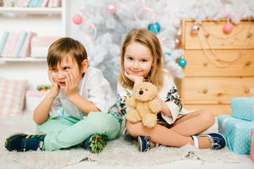 Children sitting on floor on carpet with new year decorations and christmas tree. Emotional kids faces. Handsome young boy and little princess girl looking at camera. Happy people together on holidays